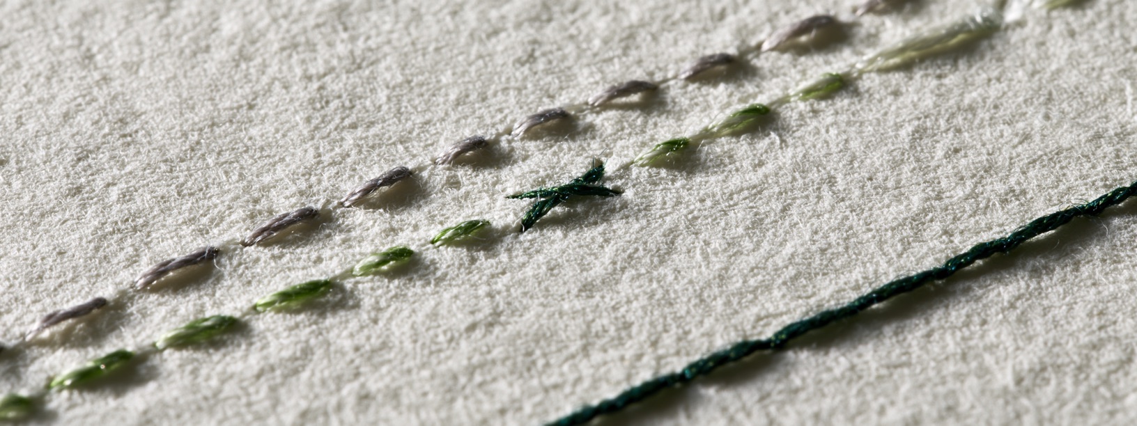 Two stitched thread lines, one graphite and one green, crossing once on handmade paper